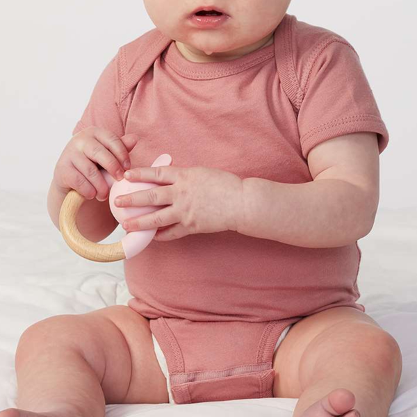 A baby wearing a dusty rose short-sleeve bodysuit sits on a white surface, holding a pink teething toy with a wooden ring.